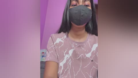 A video of a young woman with medium brown skin, wearing a gray face mask, gray shirt with white pattern, and long black hair. The background features a pink wall and a white bottle.