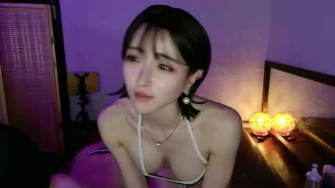 Video of a young Asian woman with fair skin, black hair, and large breasts in a low-cut top, sitting on a bed with a wooden headboard, lit by dim, purple-toned lights.