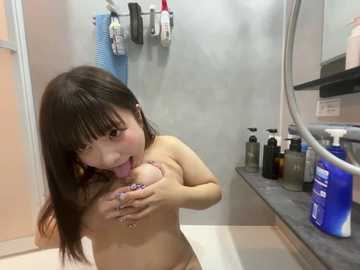 Video of a nude Asian woman with dark hair, large breasts, and flushed face licking her right nipple, in a modern bathroom with tiled walls, toiletries, and a mirror.