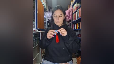 Video of a young woman with long dark hair, dressed in a black hoodie and jeans, carefully tying a red tassel, in a cluttered, dimly lit storage room with shelves filled with colorful bags.