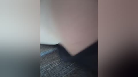 A blurred video showing a close-up of a person's torso in a dark outfit, standing in front of a beige wall, with a textured grey floor beneath. The image is out of focus, creating a dreamy, abstract effect.