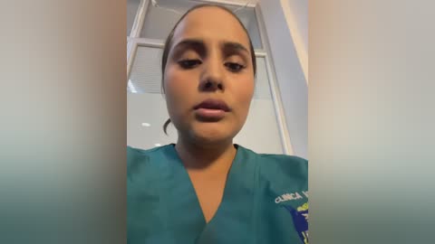 A close-up video of a woman with medium brown skin and dark hair tied back, wearing teal medical scrubs. The background features a white window frame and part of a room with a neutral color scheme.