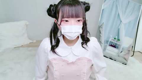 Video of a young Asian woman with long black pigtails, wearing a white face mask, pink dress, and white apron, kneeling on a white bed, surrounded by soft, pastel-colored decor.