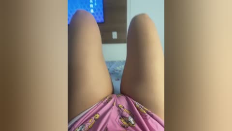 Video of a person lying on their back, legs slightly apart, wearing pink shorts with cartoon characters. Background shows a TV screen displaying a blue light and a white wall.