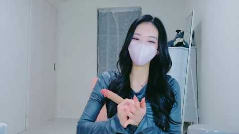 Video of an Asian woman with long black hair, wearing a white mask, denim jacket, and holding a large pink dildo in a minimalist, brightly-lit white room.