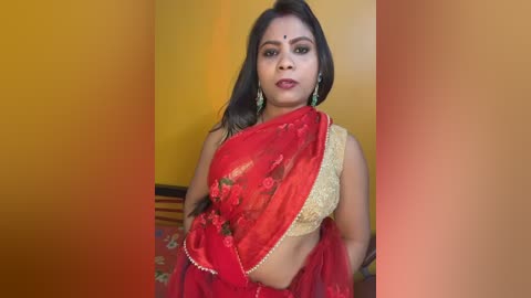 Video of a South Asian woman in a red sari with gold embroidery, holding a dupatta, wearing green earrings, against a mustard-yellow wall.