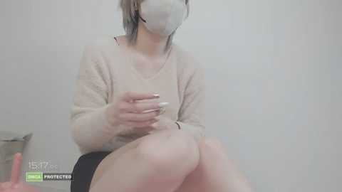 Media: Video of a young Asian woman in a beige sweater, seated in a chair, wearing a white mask, with a gray wall background.
