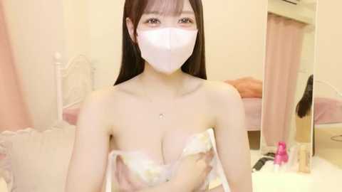 Video of an East Asian woman with long black hair, wearing a white face mask, floral bra, and sitting in a pastel-colored bedroom with pink curtains and a vanity mirror.