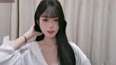Video of a young East Asian woman with long, straight black hair, wearing a white robe, in a bathroom with beige curtains. She appears relaxed, one hand in her hair.