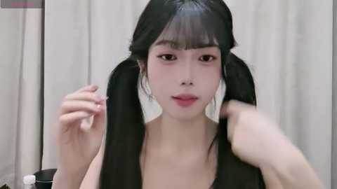 Video of an Asian woman with straight black hair styled in pigtails, light skin, wearing a black top. She's adjusting her hair, with a neutral expression. Background is a plain, light-colored curtain.