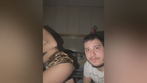 Video of a man with short hair and a beard, wearing a white t-shirt, looking at a woman in leopard print underwear, in a dimly lit kitchen with white cabinets and a stuffed animal.