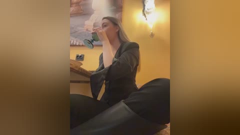 Video of a woman with long brown hair, wearing a black top and pants, sitting cross-legged on a bed in a warm-toned room with a wall-mounted lamp and a framed picture in the background.