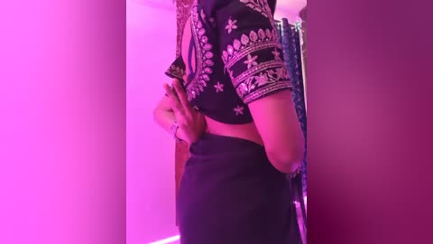 Video of a woman's midsection in a black, intricately embroidered blouse and high-waisted black skirt. The background features a pink-lit room with a partially visible curtain.
