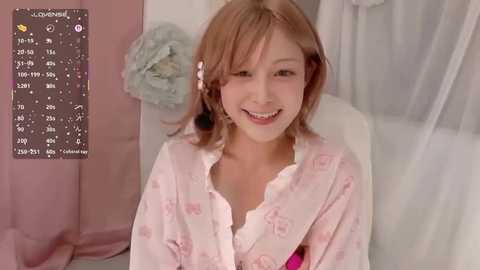 A video of a smiling young Asian woman with light brown hair in a pastel floral robe, sitting in a softly lit room with a pink curtain and white drapes.