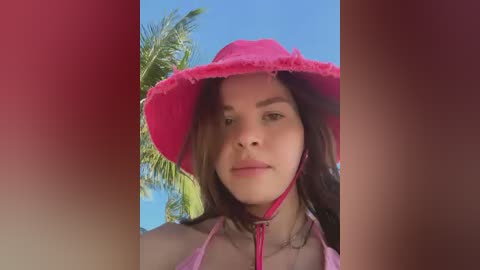 Video of a young woman with light skin, brown hair, and a pink hat, standing outdoors against a clear blue sky with a palm tree in the background. She wears a pink bikini top and has a neutral expression.