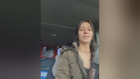 Video of a young woman with long dark hair, wearing a green puffer jacket and a black top, standing in a car with a red emergency light in the background.
