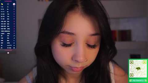 Video of a young Asian woman with long black hair, wearing a white top, looking downward with closed eyes. Background shows a blurred room with a TV displaying a website and a QR code.