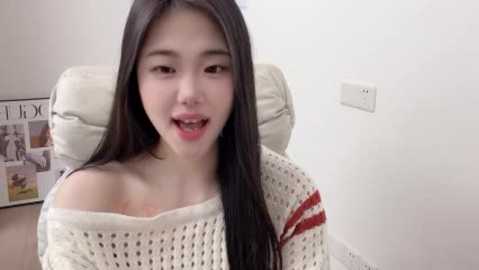 Video of an East Asian woman with long black hair, wearing a white off-shoulder knitted sweater, sitting on a beige chair in a minimalist room.