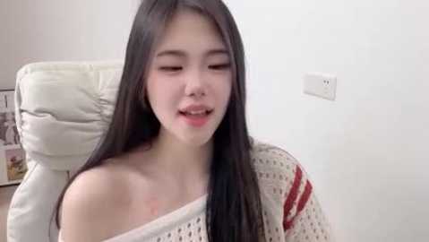 A young Asian woman with long black hair, fair skin, and a slender build, wearing a white, off-shoulder knit sweater, sits in a white chair, eyes closed, smiling softly.