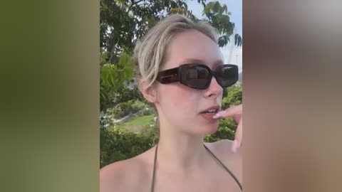 Video of a fair-skinned woman with short blonde hair, wearing dark sunglasses and a black bikini top, biting her finger in a natural outdoor setting with lush greenery and clear skies.