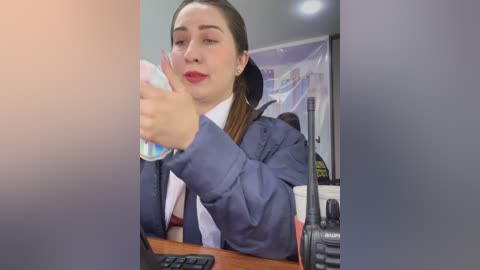 A video of a young woman with light skin, brown hair, and red lipstick, wearing a blue blazer over a white shirt, drinking from a colorful water bottle at a desk, with a microphone and papers in the background.