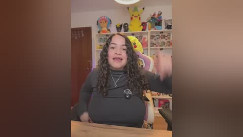 Video of a plus-size woman with curly hair, wearing a black top and Pikachu plush toy, smiling while holding a camera in a brightly decorated room filled with Pok\u00e9mon figurines and Pikachu-themed decor.