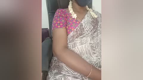 Video of a person wearing a pink blouse with a floral pattern, a white saree with a palm leaf design, and a white flower garland. The background shows a dark chair and a blurred purple cushion.