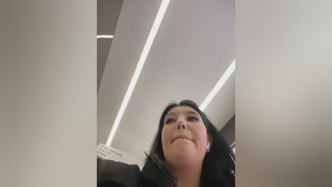 Video of a smiling woman with long black hair, wearing a black jacket, standing in a modern, well-lit room with white walls and recessed lighting.