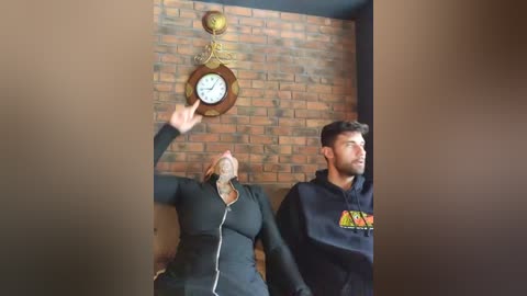 Video of a woman in a black zip-up hoodie, hands raised, looking up at a clock on a brick wall, next to a man in a black hoodie with colorful graphic, both appearing confused.