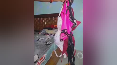 Video of a woman holding a pink and black lingerie set in a cluttered bedroom with a bed, nightstand, and colorful bedding.