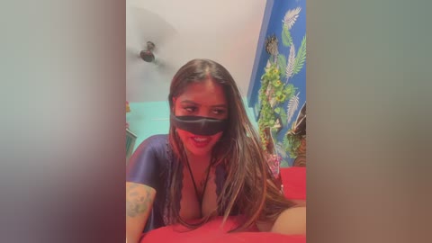 Video of a smiling woman with long brown hair, wearing a black mask and a blue top, lying on a red bed, in a room with a blue wall featuring a palm leaf design.