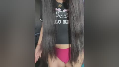 Video of a slender, light-skinned person with long, straight, dark hair. They wear a black cropped shirt with \"LOL KITTIES\" and a necklace, and bright pink panties. Background is blurred.