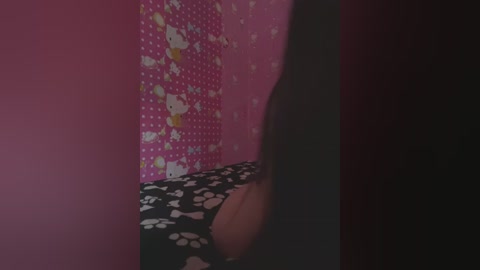 Video of a person's bare back against a pink wall with Hello Kitty-themed wallpaper. The individual is wearing black pants with white paw print patterns. The room is dimly lit, creating a mysterious atmosphere.