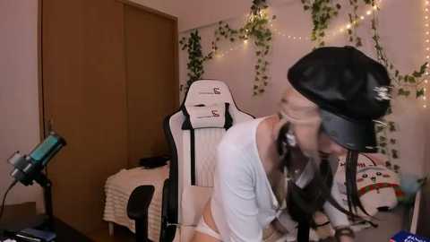Video of a young woman in a white, off-the-shoulder top and black beret, leaning over a desk with a gaming chair in a cozy room with fairy lights and a plush toy.