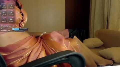 A video of a person in a VR headset, wearing a pink robe, reclining on a beige couch, with a dark chair in the foreground. The room has beige walls and curtains.