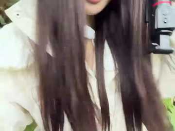 Video of a young woman with long, wavy, dark brown hair, partially cropped, wearing a white shirt. Background features a blurred, beige wall and a black shelf with a silver bottle.
