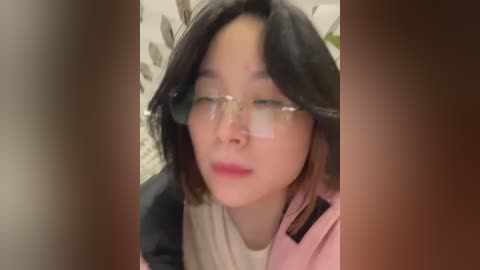 Video of an Asian woman with shoulder-length black hair, wearing glasses and a pink jacket, with a blurred background of a white wall and patterned wallpaper.