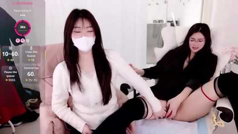 Video of two Asian women, one in a white mask and sweater, the other in a black dress and thigh-high socks, sitting on a pink sofa in a modern living room.