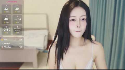 Video of an East Asian woman with long black hair, fair skin, and a slim figure, wearing a white lace bra. She stands in a hospital room with a bed and blue curtains in the background. A health tracker screen shows her vitals.