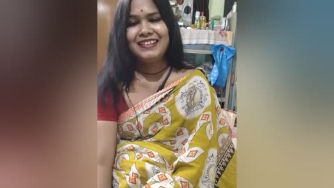 Video of a smiling South Asian woman with medium skin tone, wearing a yellow sari adorned with white circular designs, in a cluttered, possibly kitchen-like setting.