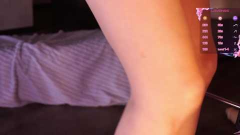 Video of a nude, fair-skinned person's leg standing beside a bed with a rumpled gray sheet and a TV displaying a stock market chart. The background is dimly lit, creating a warm, intimate atmosphere.