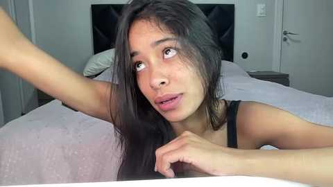 Media: Video of a young woman with long, dark hair, light skin, and a slim physique, lying on a bed with a white blanket, wearing a black tank top. She has a contemplative expression, with her arm raised.