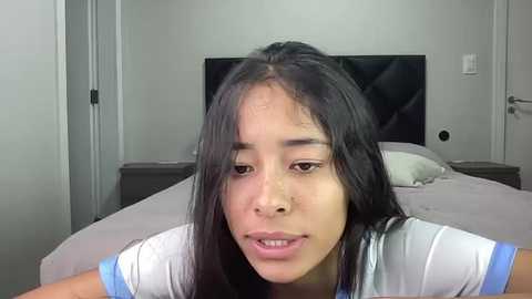 Video of a young Asian woman with long black hair, wearing a white shirt, in a modern, minimalist bedroom with a gray bed and flat-screen TV.