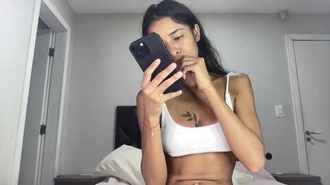 Media: A video of a slender, light-skinned woman with long black hair, wearing a white crop top, taking a mirror selfie in a minimalist bedroom with white walls and a TV.