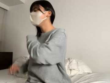 A video of a person wearing a white mask and gray sweatshirt, standing in a dimly lit room with white walls and a bed covered in white sheets.