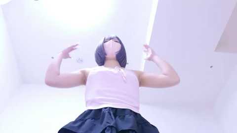 Video of a young woman with a short bob haircut, wearing a light pink tank top and a black pleated skirt, standing in a brightly lit room with white walls.