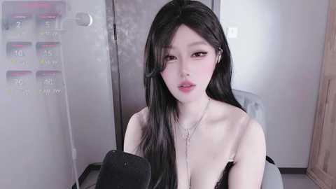 Video of an East Asian woman with long black hair and pale skin, wearing a black lace bra, sitting in a clinical room with a temperature monitor and medical equipment.
