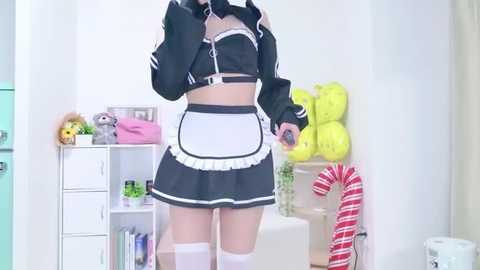Video of a woman in a black and white maid outfit, standing in a bright, whimsical room with a white bookshelf, green and yellow decorations, and a red and white candy cane.