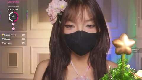 Video of an Asian woman with long black hair adorned with a pink flower, wearing a black face mask. She holds a Christmas tree with a star.