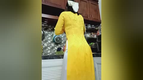Video of a woman in a vibrant yellow dress with a high slit, standing in a modern kitchen with wooden cabinets, a patterned backsplash, and various kitchen items on shelves.